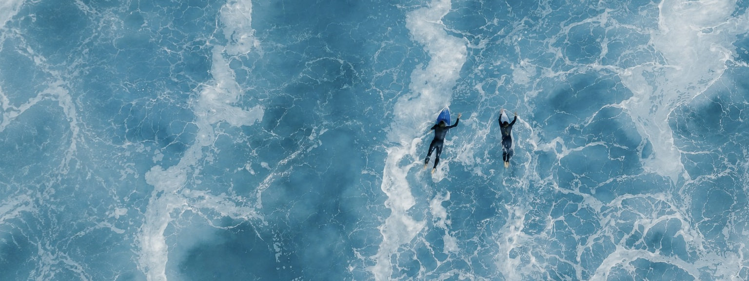 Two surfers paddling through blue ocean foam during the KANOA End of Summer Sale